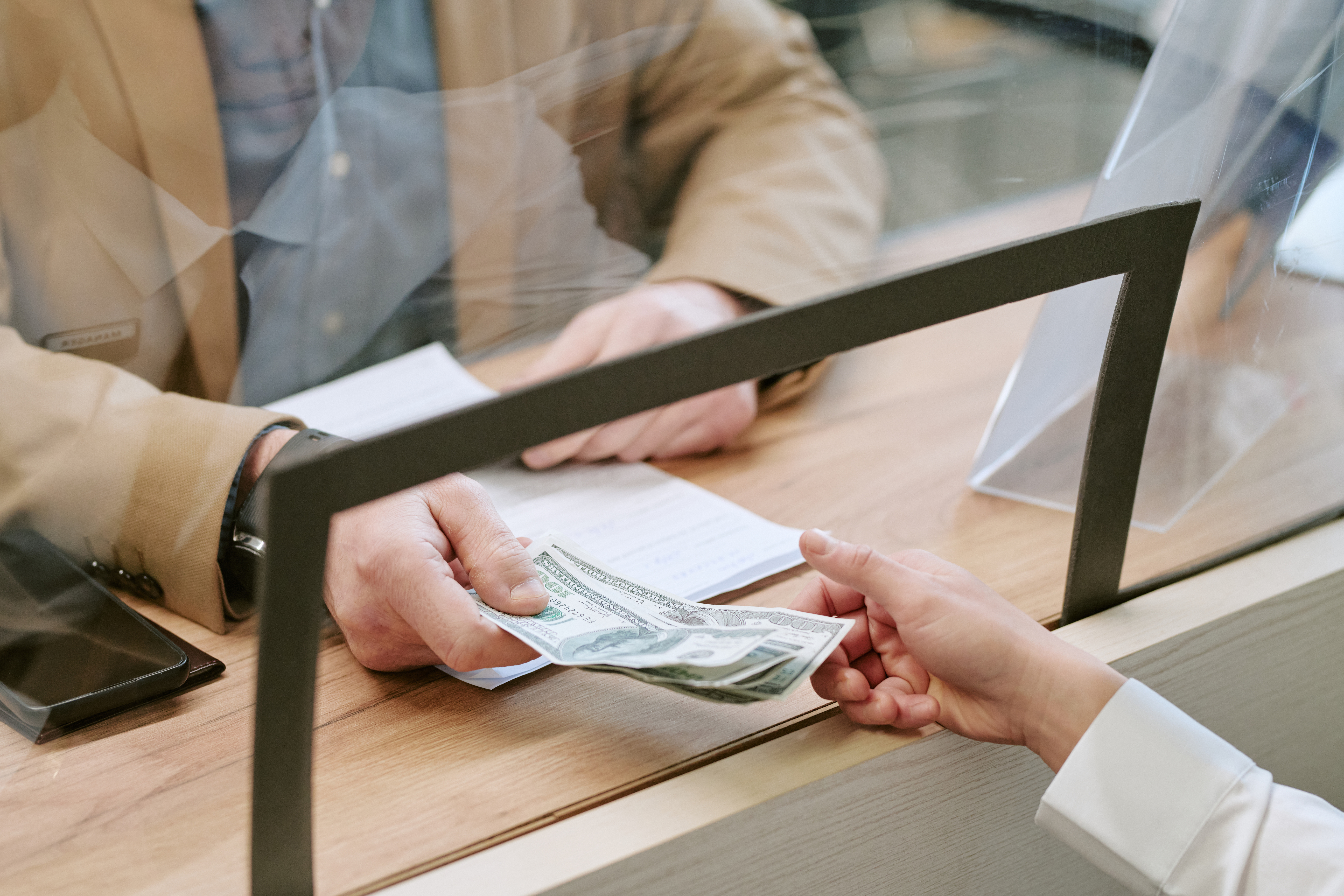 Caucasian man handing cash to bank teller across counter, both hands visible through protective glass, financial transaction occurring inside bank branch setting